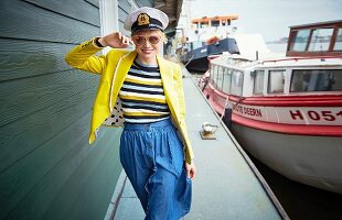 A blonde woman wearing a yellow blazer, a striped jumper, a denim skirt and a captain's hat