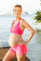 A pregnant woman on a jetty wearing a pink sports bra and shorts