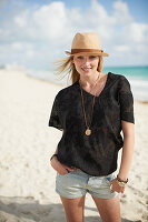 A blonde woman on a beach wearing a brown blouse, shorts and a hat
