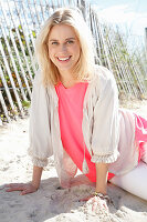 Junge blonde Frau in weißer Bluse und rosa Unterziehshirt am Strand sitzend