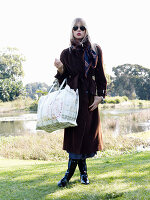 A young blonde woman standing in a meadow wearing sunglasses and a brown coat