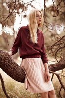 A young blonde woman leaning against a tree wearing a dark red jacket and a pink pleated skirt