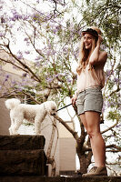 Junge Frau mit Hut in hellem Top und Shorts auf Steinmauer mit Hund