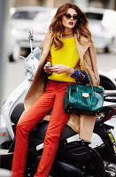 A young woman wearing a yellow jumper, red trousers and a sand-coloured coat