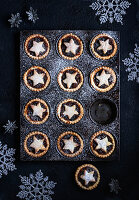 A tray of mince pies decorated with stars dusted with icing sugar on a dark background with Christmas decorations