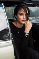 A young, dark-haired woman wearing a black dress sitting in a car