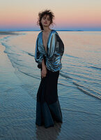 A dark-haired woman wearing a long evening dress by the sea at sunset