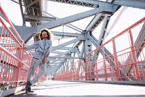 Dunkelhaarige Frau mit Sonnenbrille in Jeans-Jumpsuit auf einer Brücke
