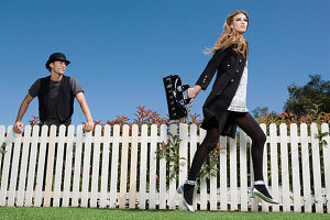 A young woman wearing a black coat and black tights with a man wearing a black hat leaning on a fence