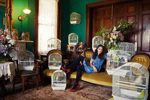 A dark-haired woman sitting on a sofa in a living room surrounded by birdcages