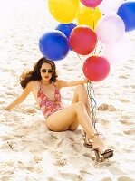 A brunette woman sitting on a beach wearing a bathing suit and holding balloons