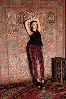 A young woman wearing a black top and dark-red trousers in front of a hanging rug