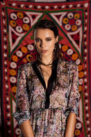 A young woman wearing a patterned dress standing against a hanging rug