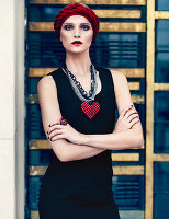 A young woman wearing a black cocktail dress, a red turban headband and a necklace with a heart-shaped pendant