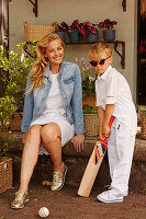 A little boy wearing a white outfit holding a cricket bat with his mother wearing a white dress and denim jacket