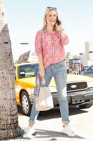 A blonde woman wearing jeans and a floral blouse on the phone outside