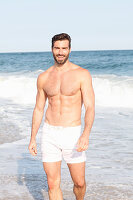 A young topless man walking along a beach wearing shorts