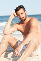 Junger Mann mit Shorts und nacktem Oberkörper sitzt am Strand