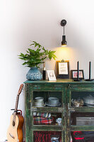 Vase of leafy branches, pictures and candles on top of green cabinet below wall-mounted lamp