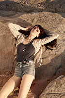 A brunette woman wearing a brown top, a jacket and shorts lying on rocks