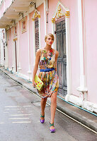 A young woman wearing a colourful, patterned summer dress with purple sandals and a clutch bag