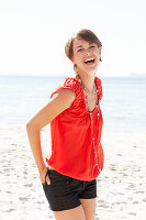 A mature brunette woman on a beach wearing a red shirt, a necklace and black shorts