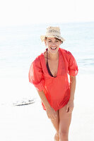 A mature brunette woman on a beach wearing a red shirt, a black bathing suit and a beige hat