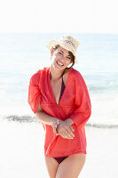 Reife brünette Frau mit rotem Shirt, schwarzem Badenanzug und beigem Hut am Strand