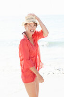 A mature brunette woman on a beach wearing a red shirt and a beige hat