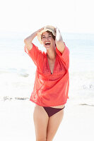 A mature brunette woman on a beach wearing a red shirt, a black bathing suit and a beige hat