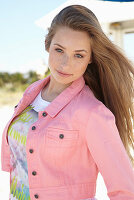 A young blonde woman on a beach wearing a printed t-shirt and a pink denim jacket