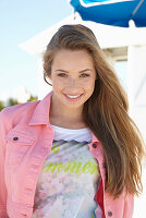 A young blonde woman on a beach wearing a printed t-shirt and a pink denim jacket