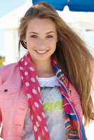 A young blonde woman on a beach wearing a printed t-shirt, a pink denim jacket and a patterned scarf