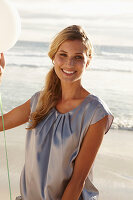 A mature blonde woman on a beach wearing a silver summer dress and holding a white balloon