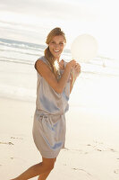A mature blonde woman on a beach wearing a silver summer dress and holding a white balloon