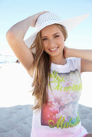 Junge blonde Frau mit buntem Shirt, weißem Sommerhut am Strand