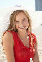 Junge blonde Frau in rotem Sommerkleid mit weißem Hut am Strand