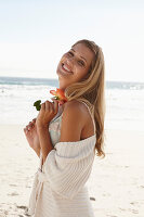 Reife blonde Frau in Dessous und Strickjacke und mit Blume in der Hand am Strand