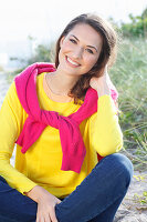 A brunette woman wearing a yellow jumper and jeans with a pink jumper over her shoulders