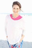 Brünette Frau in pinkfarbenem T-Shirt, weißem Pulli und bunter Hose am Strand