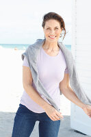 Brunette woman wearing lilac T-shirt, jeans and grey sweater over shoulders on beach