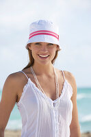 A young brunette woman on a beach wearing a white top and a white hat