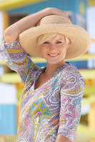 A mature blonde woman with short hair on a beach wearing a patterned summer dress and a hat