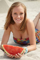 A young blonde woman on a beach wearing a colourful summer dress holding a wedge of melon