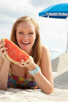 Junge blonde Frau im bunten Sommerkleid hält Melone am Strand