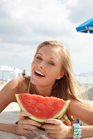 A young blonde woman on a beach wearing a colourful summer dress holding a wedge of melon