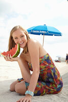 A young blonde woman on a beach wearing a colourful summer dress holding a wedge of melon