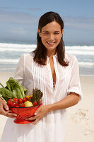 A young brunette woman on a beach wearing a white summer dress and holding a bowl of vegetables