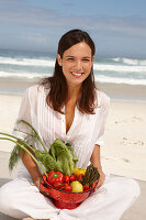 A young brunette woman on a beach wearing a white summer dress and holding a bowl of vegetables