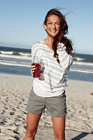Brünette Frau mit Getränk in gesteiftem Shirt und kurzer Hose am Sandstrand